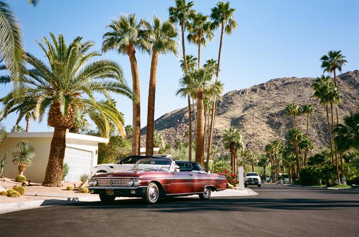 A shiny red convertible car is parked on a street lined with palm trees and desert mountains in the background.