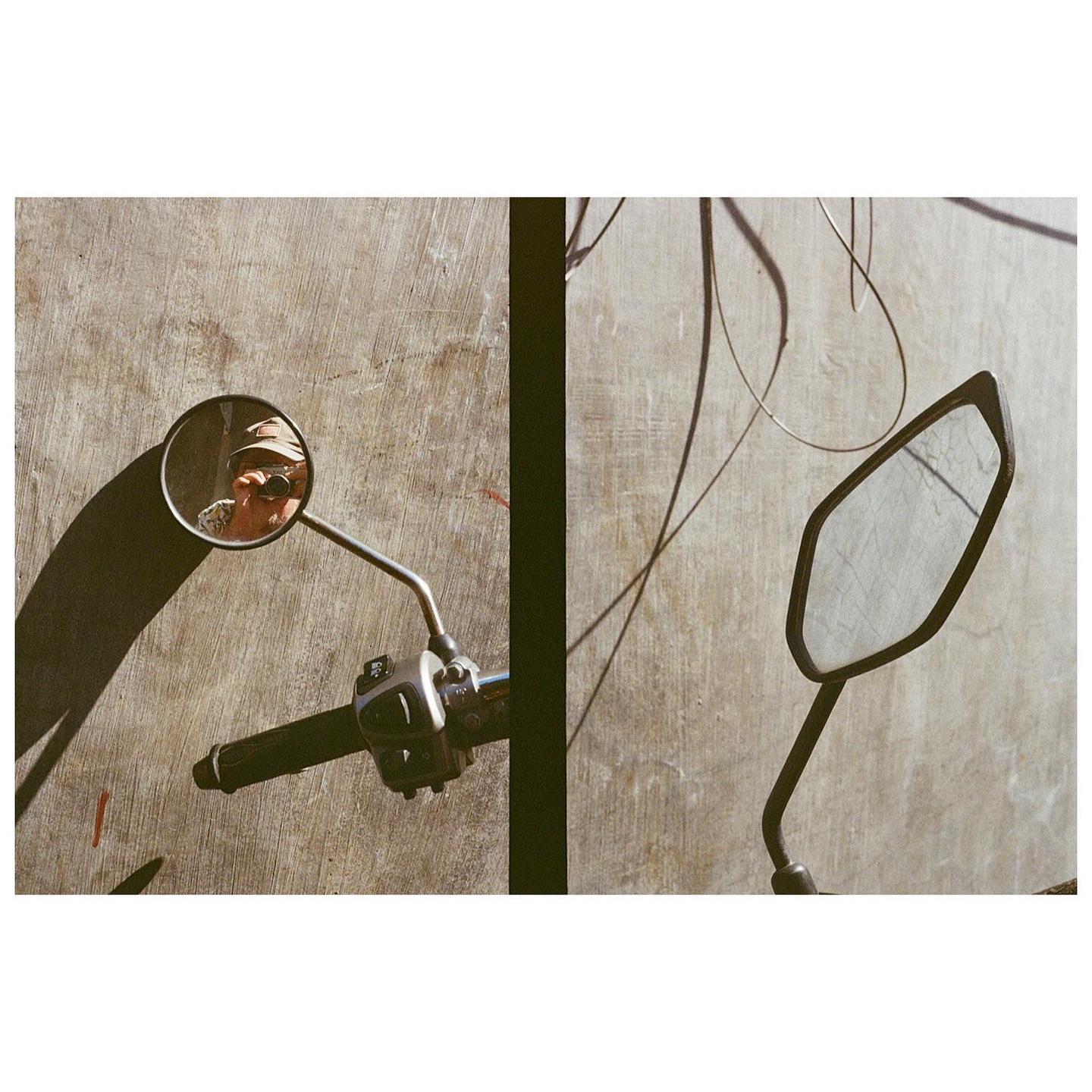 A man wearing a hat and sunglasses is reflected in a motorcycle mirror.