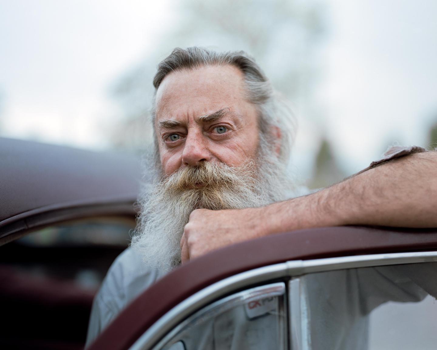 A man with a long white beard leans on a car door.