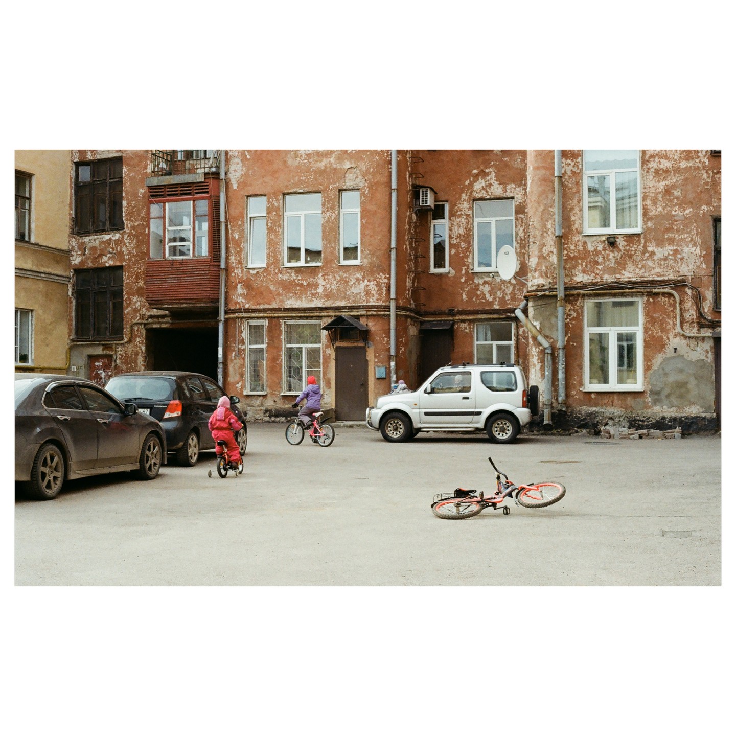 Two children ride bicycles in a paved courtyard surrounded by old buildings and parked cars.