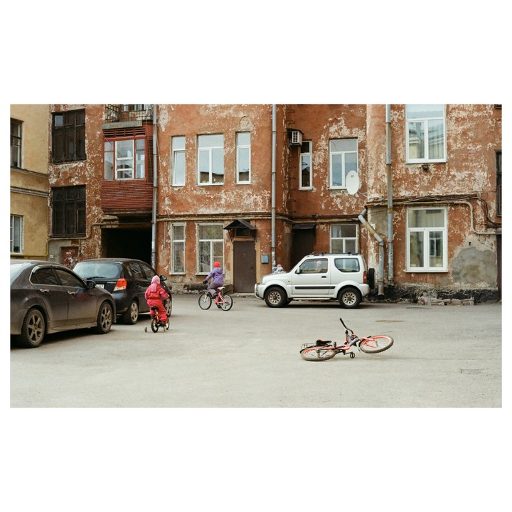 Two children ride bicycles in a paved courtyard surrounded by old buildings and parked cars.