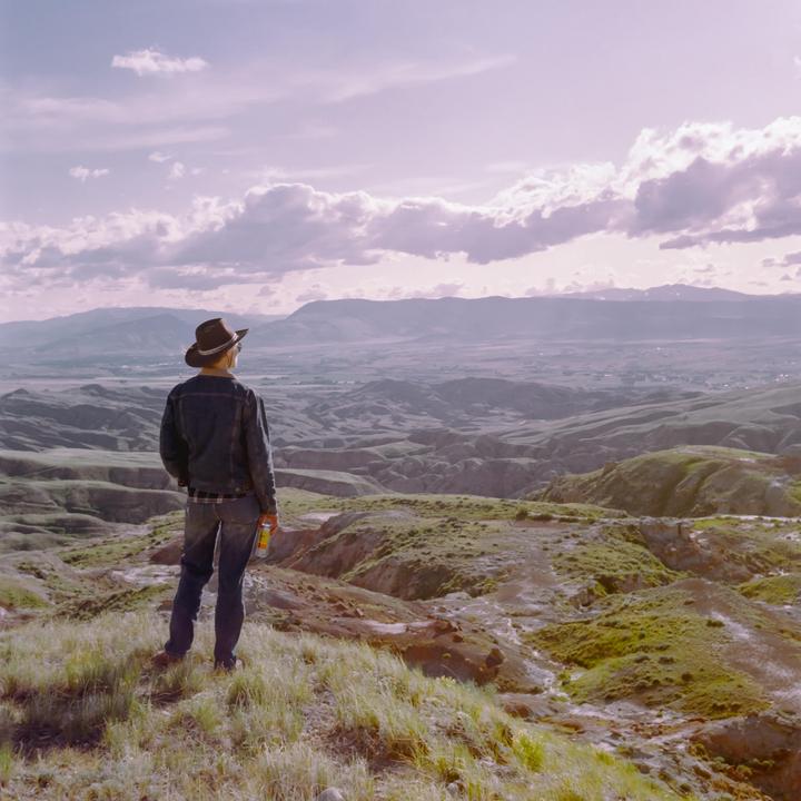 A man in a cowboy hat stands on a grassy hill overlooking a vast landscape of rolling hills and mountains under a cloudy sky.