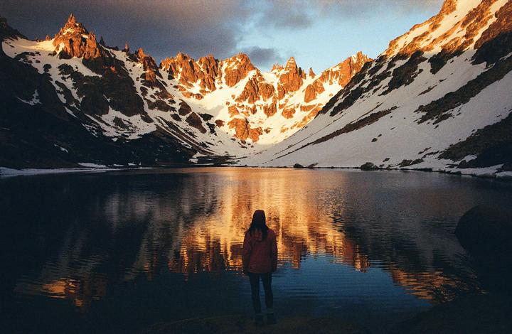 A person stands at the edge of a lake, looking at snow-capped mountains reflecting in the water at dawn.
