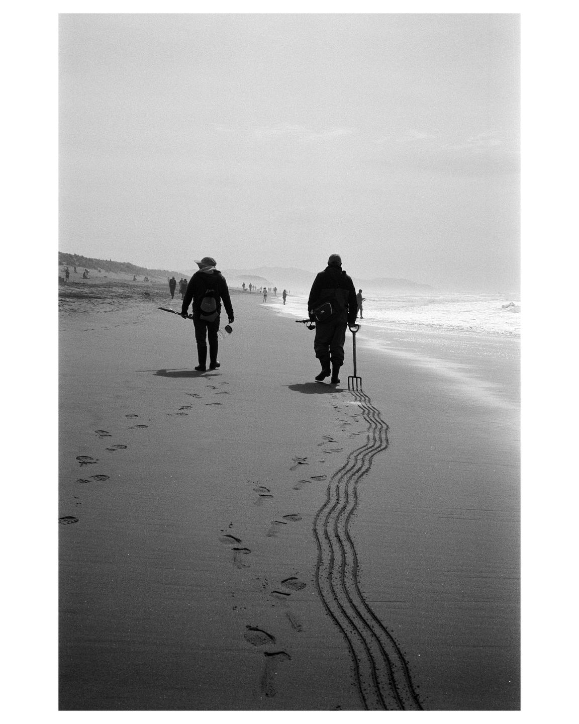 Two people walk along a beach, one dragging a rake that leaves wavy lines in the sand.