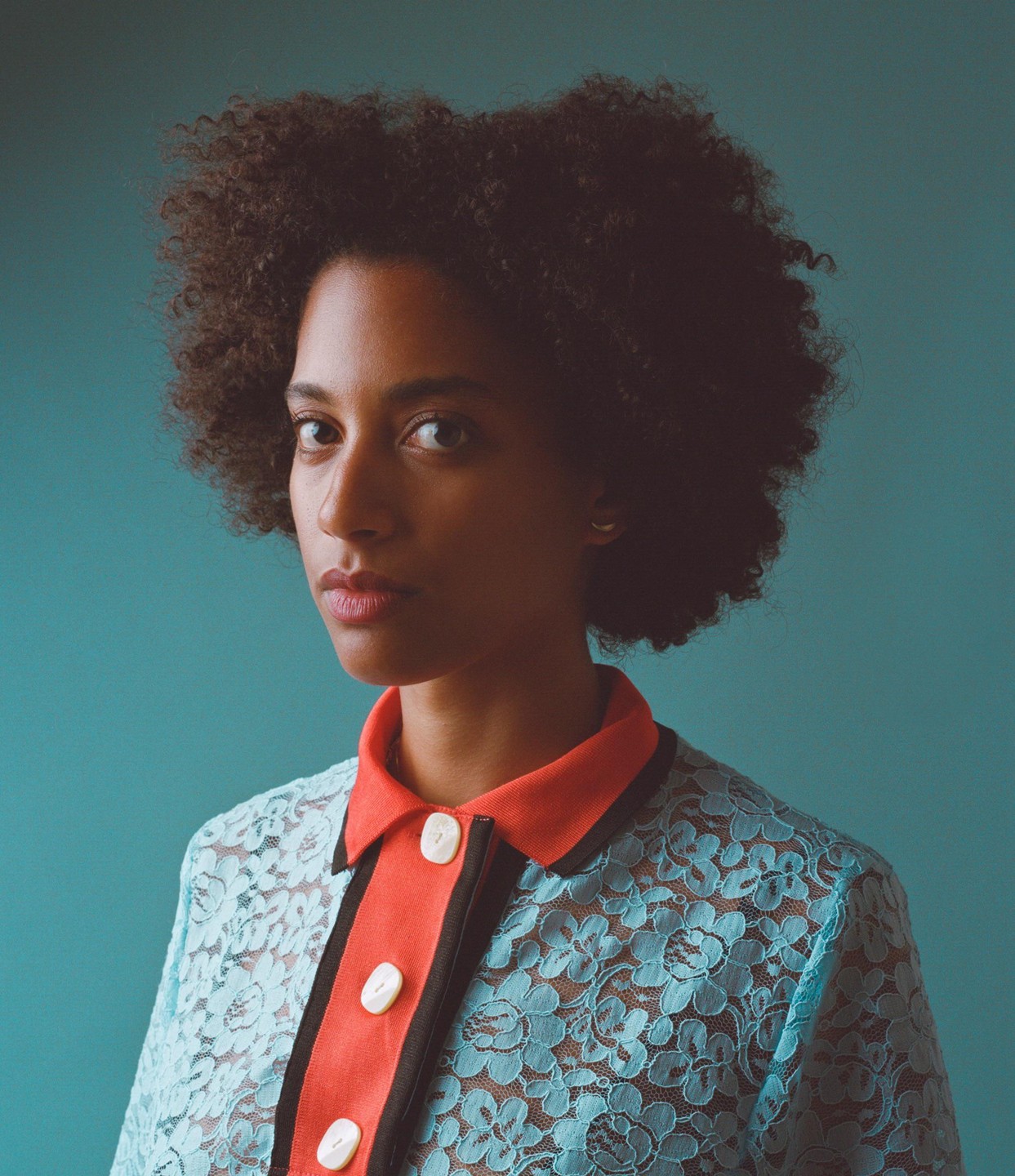 A woman with curly hair wears a blue lace shirt with an orange collar.