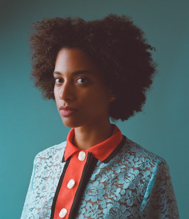 A woman with curly hair wears a blue lace shirt with an orange collar.