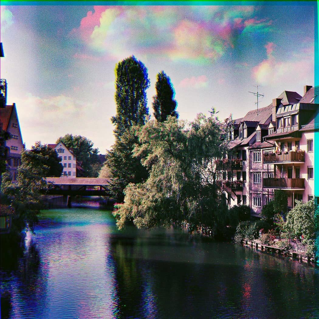 A river flows past buildings and trees under a sky with colorful clouds.