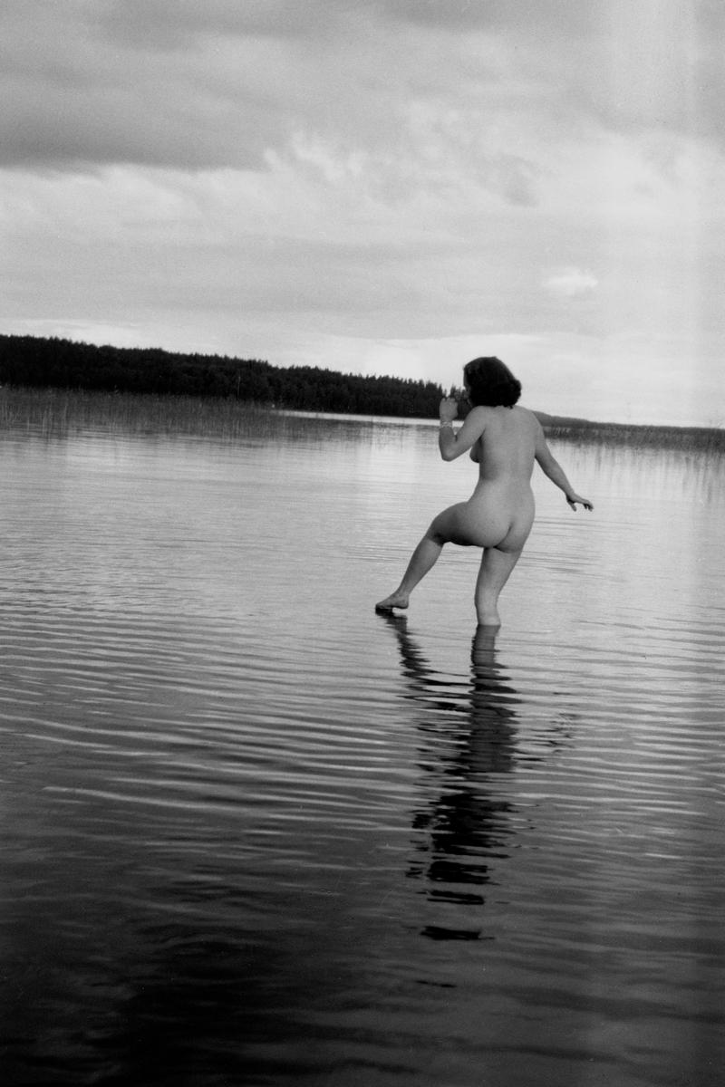 A nude woman stands in a lake with a forest in the background under a cloudy sky.