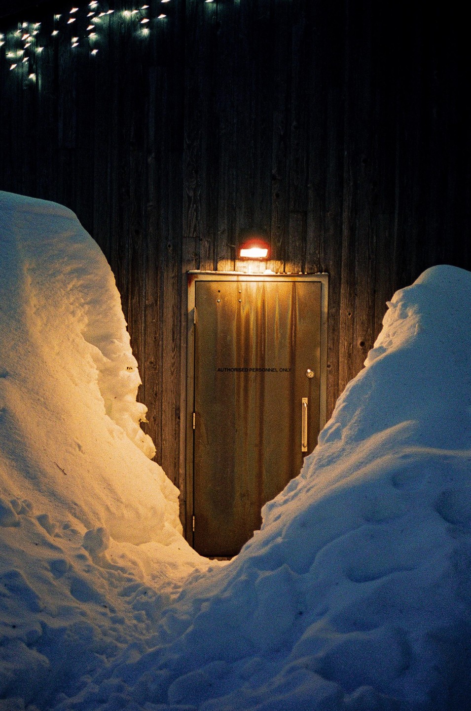 A metal door with a light above it is surrounded by snowdrifts at night.