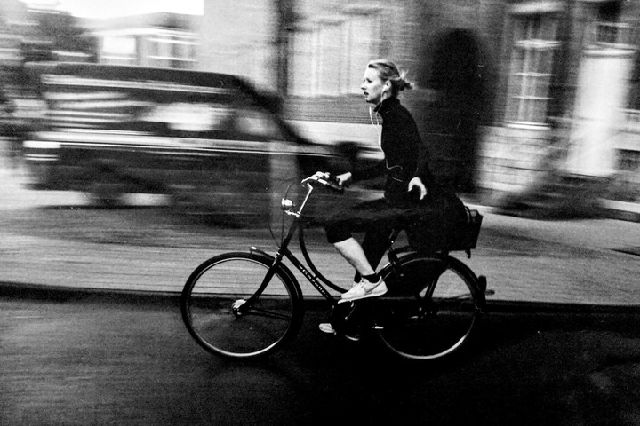A woman with earbuds rides a bicycle past a blurred vehicle on a street.