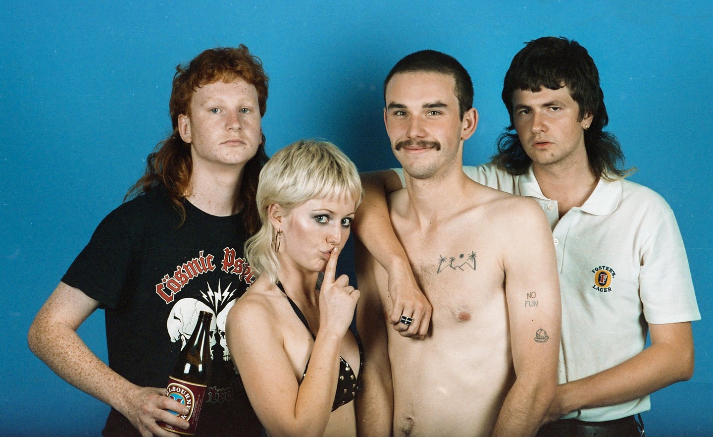Four people pose for a group portrait against a blue background, with one woman making a shushing gesture and one man holding a beer bottle.
