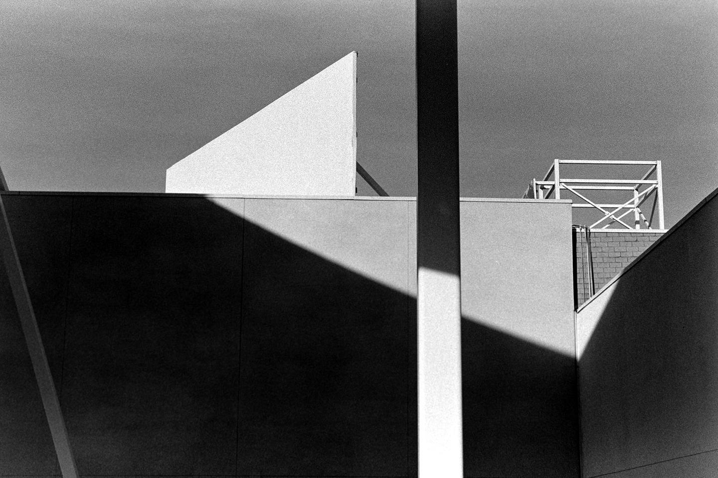 A black and white photograph shows the abstract shapes of a building with strong shadows and a metal structure on the roof.