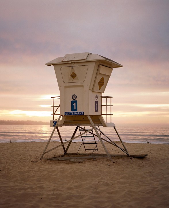 A lifeguard tower stands on a sandy beach with the ocean and a colorful sky in the background.