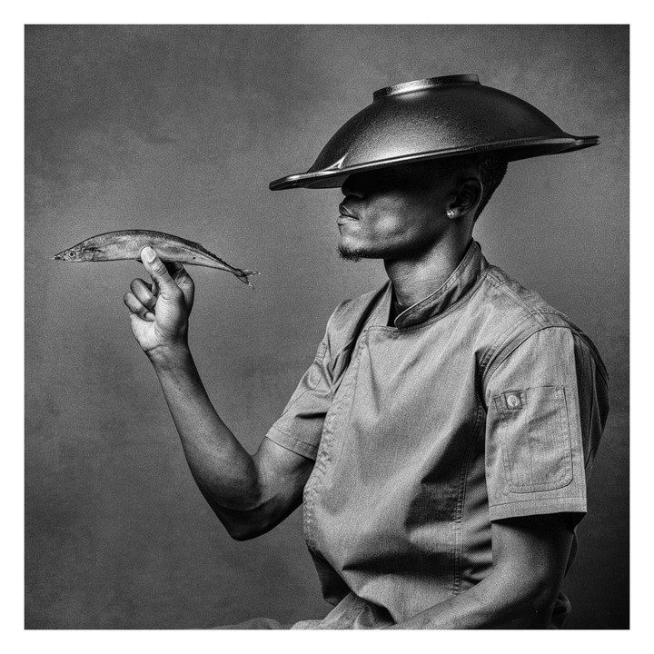 A man wearing a chef coat and a wok on his head holds a fish in his hand.