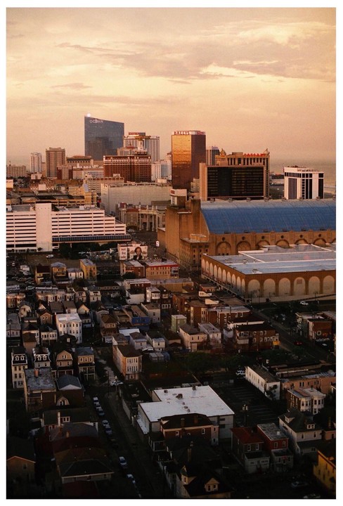 An aerial view shows a city with tall buildings and smaller houses under a cloudy sky at sunset.