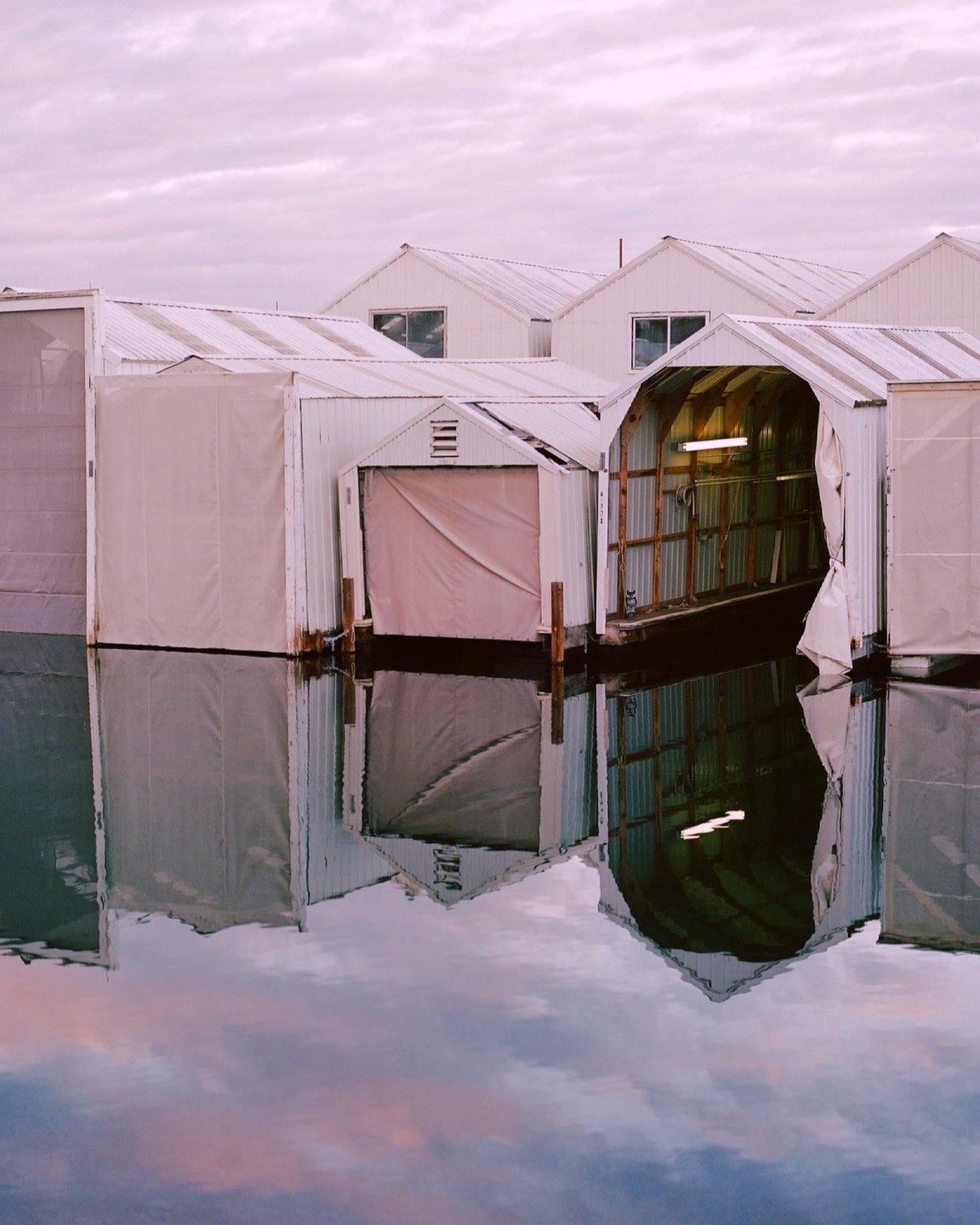 Several white boathouses with peaked roofs are reflected in the still water under a cloudy sky.