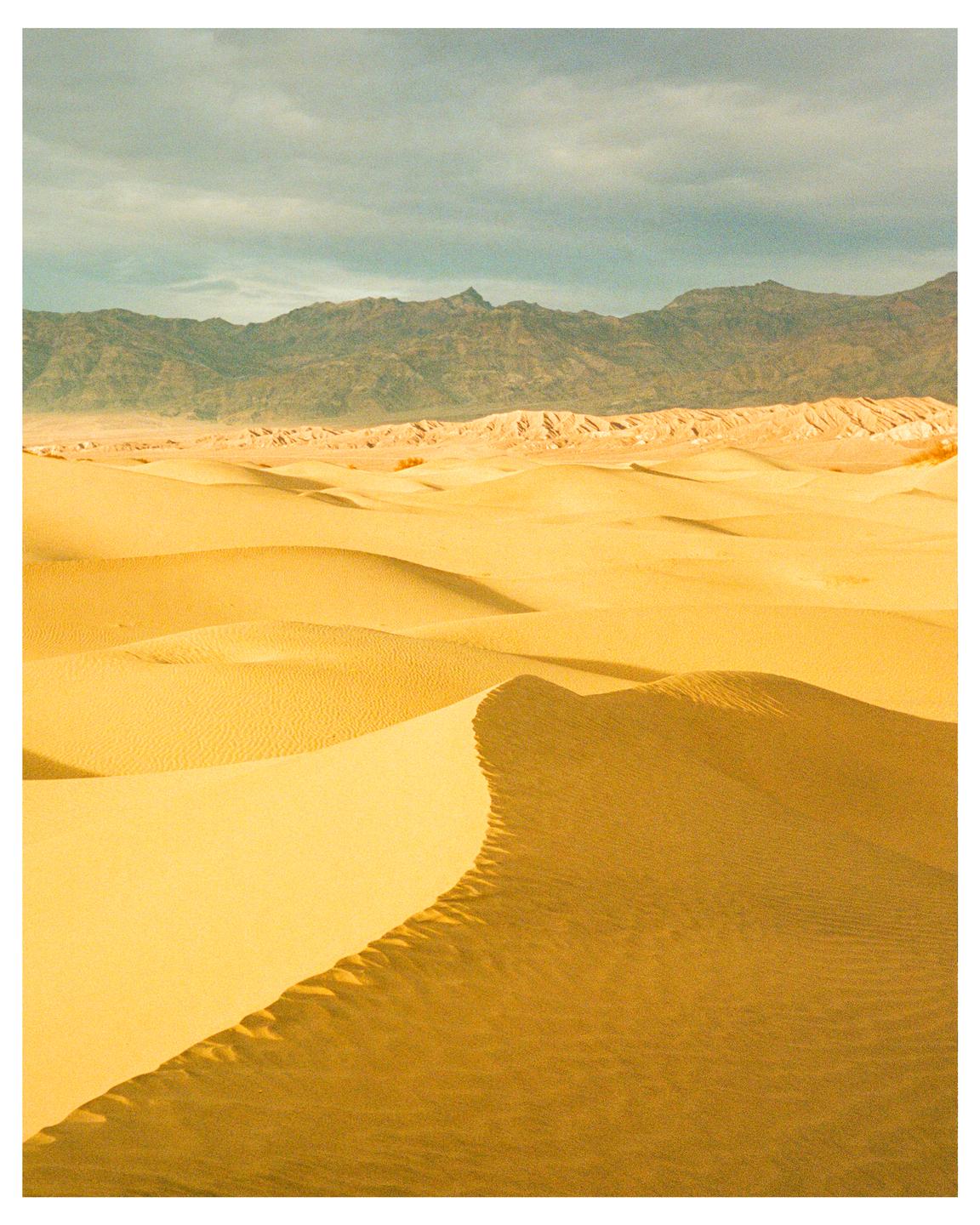 Sand dunes stretch across the desert floor with mountains in the distance under a cloudy sky.