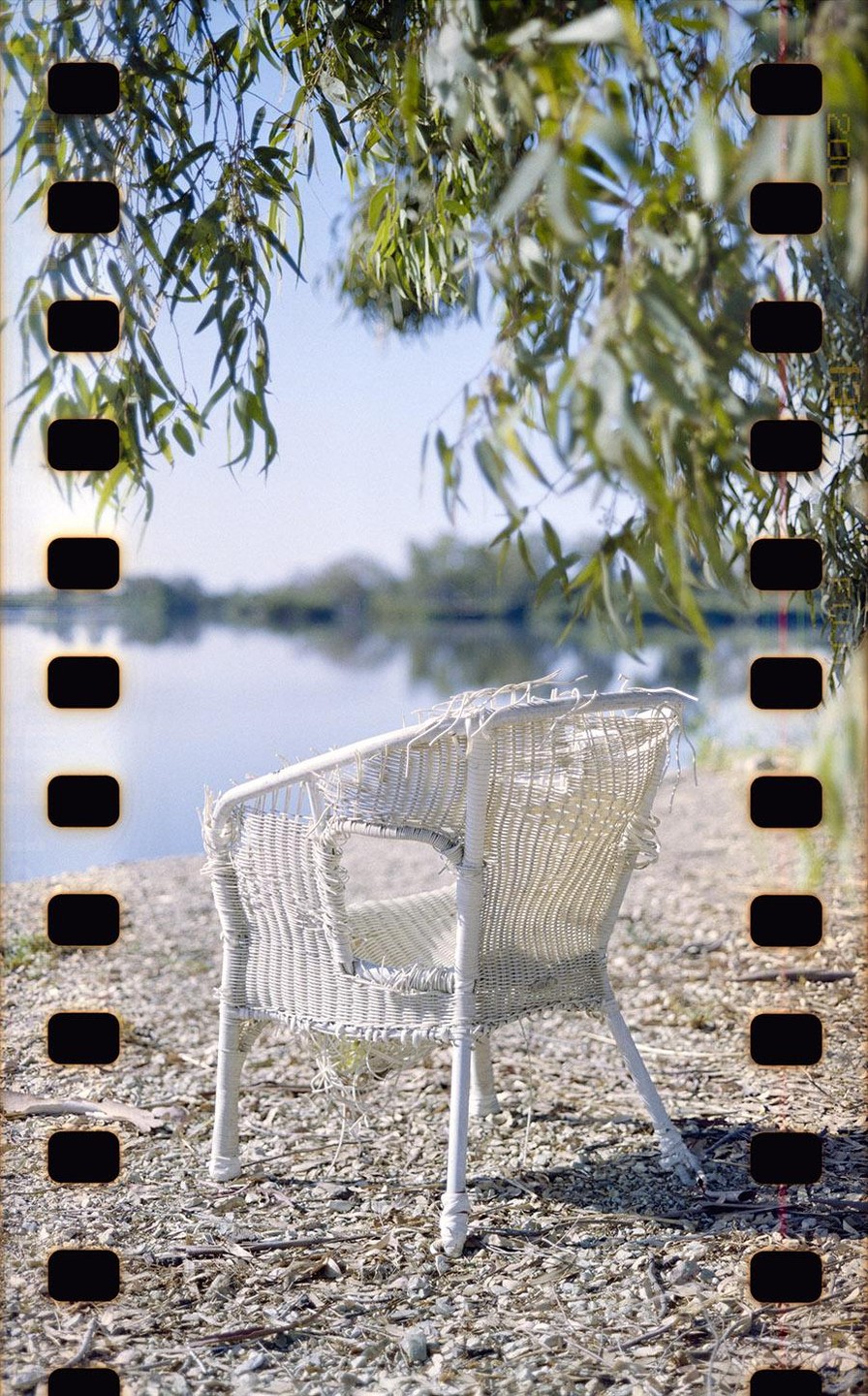 A white wicker chair sits on a rocky shore next to a body of water, with green leaves hanging from above.