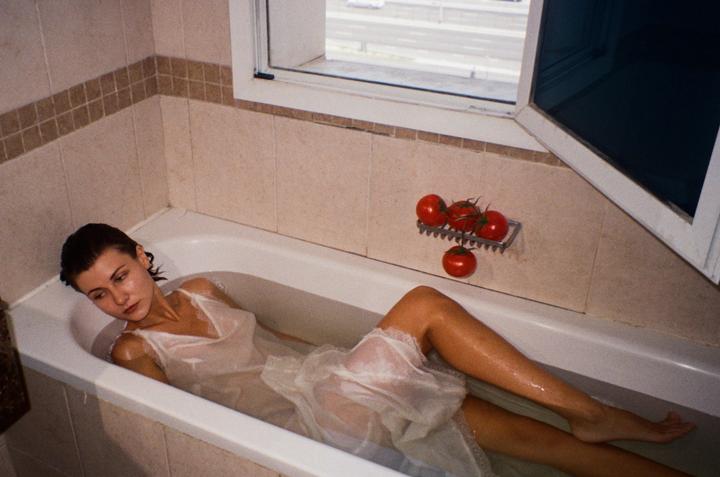 A woman in a white slip lies in a bathtub with tomatoes on a soap dish nearby.