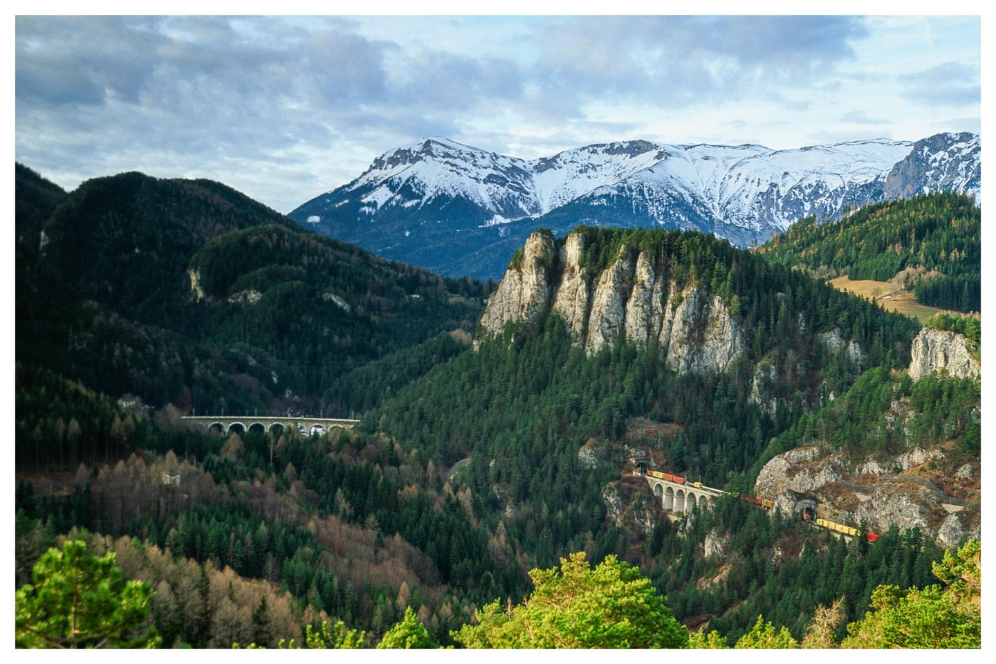A train crosses a viaduct through a valley with snow-capped mountains in the background.