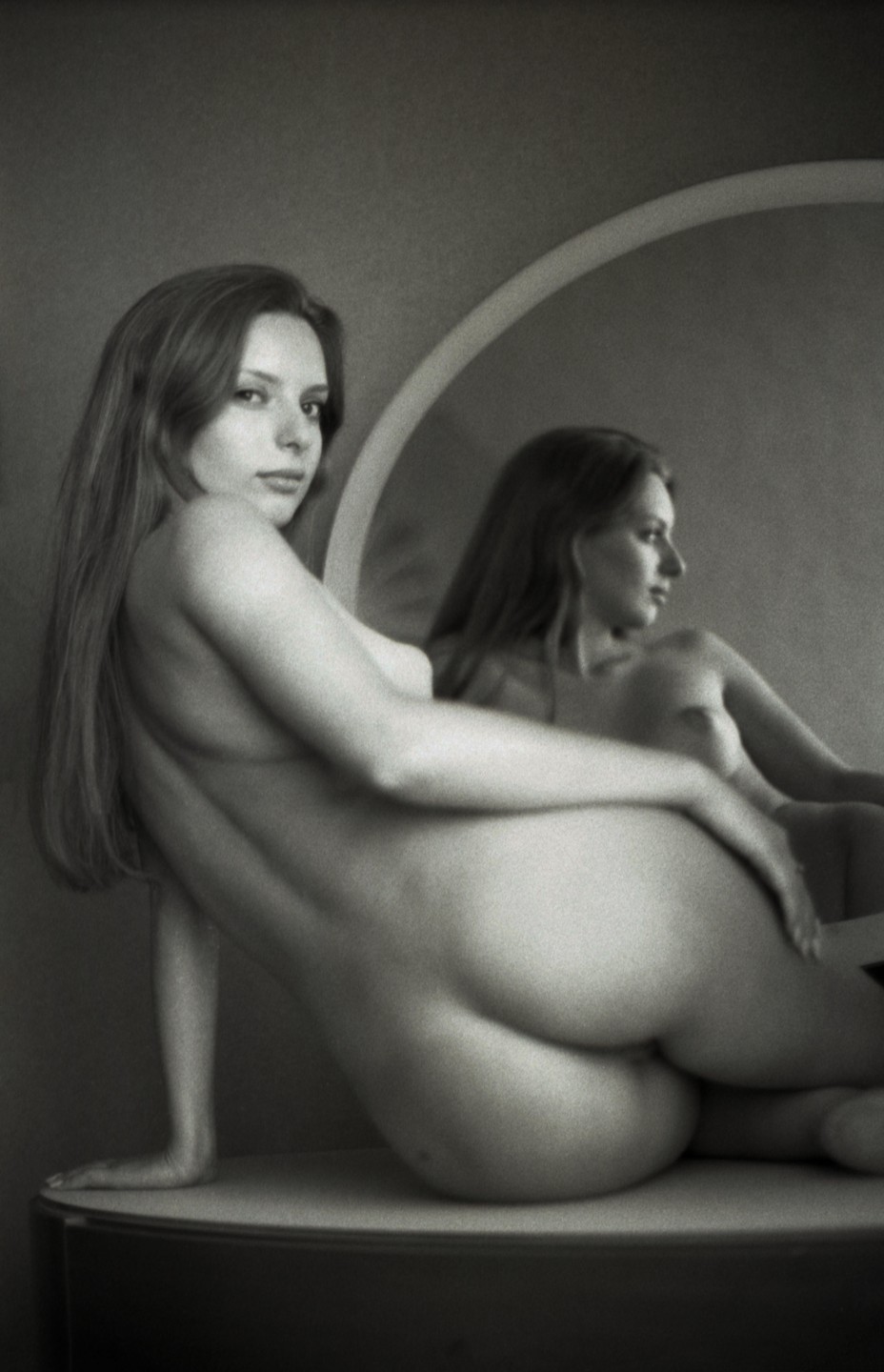 A woman is posing nude on a surface in front of a mirror, with her reflection visible in the mirror.