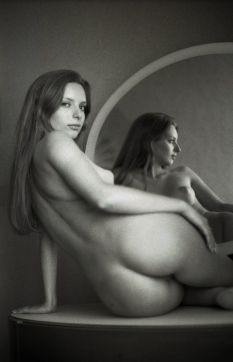 A woman is posing nude on a surface in front of a mirror, with her reflection visible in the mirror.