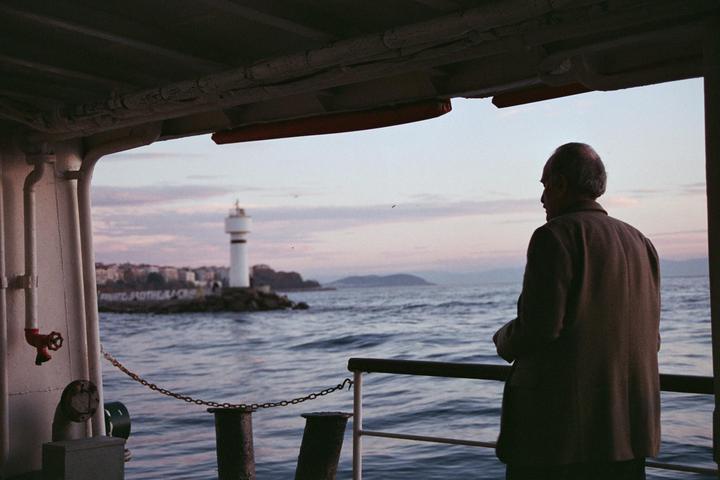 A man stands on a ferry looking out at a lighthouse and the sea at dusk.