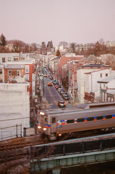 A train crosses a bridge over a street lined with cars and buildings.
