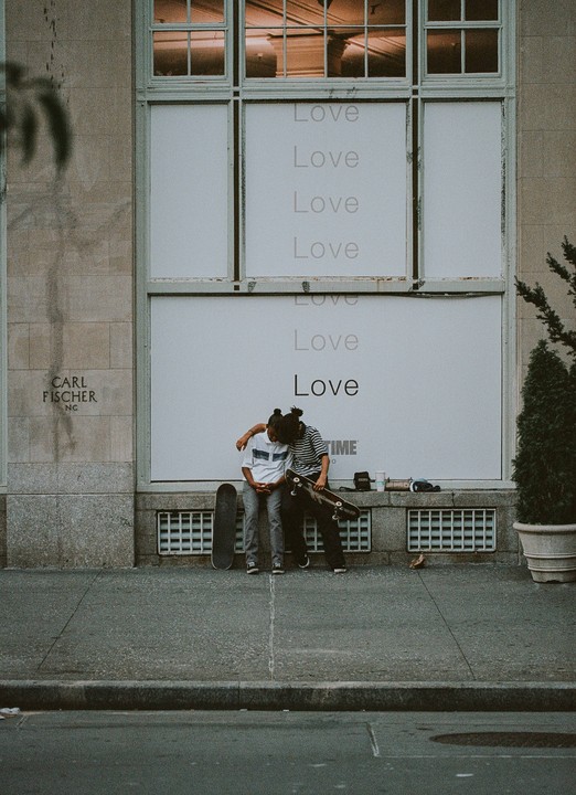 Two young men with skateboards sit on a ledge in front of a building with "Love" written on its windows.