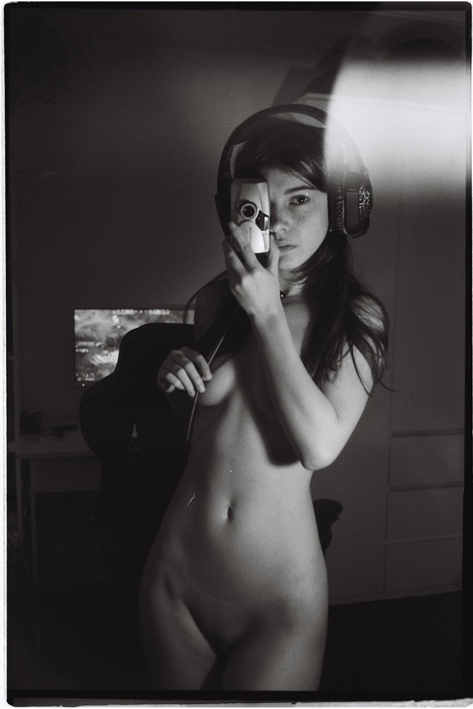 A woman wearing headphones holds a small camera to her face, obscuring one eye, while standing nude in a dimly lit room.