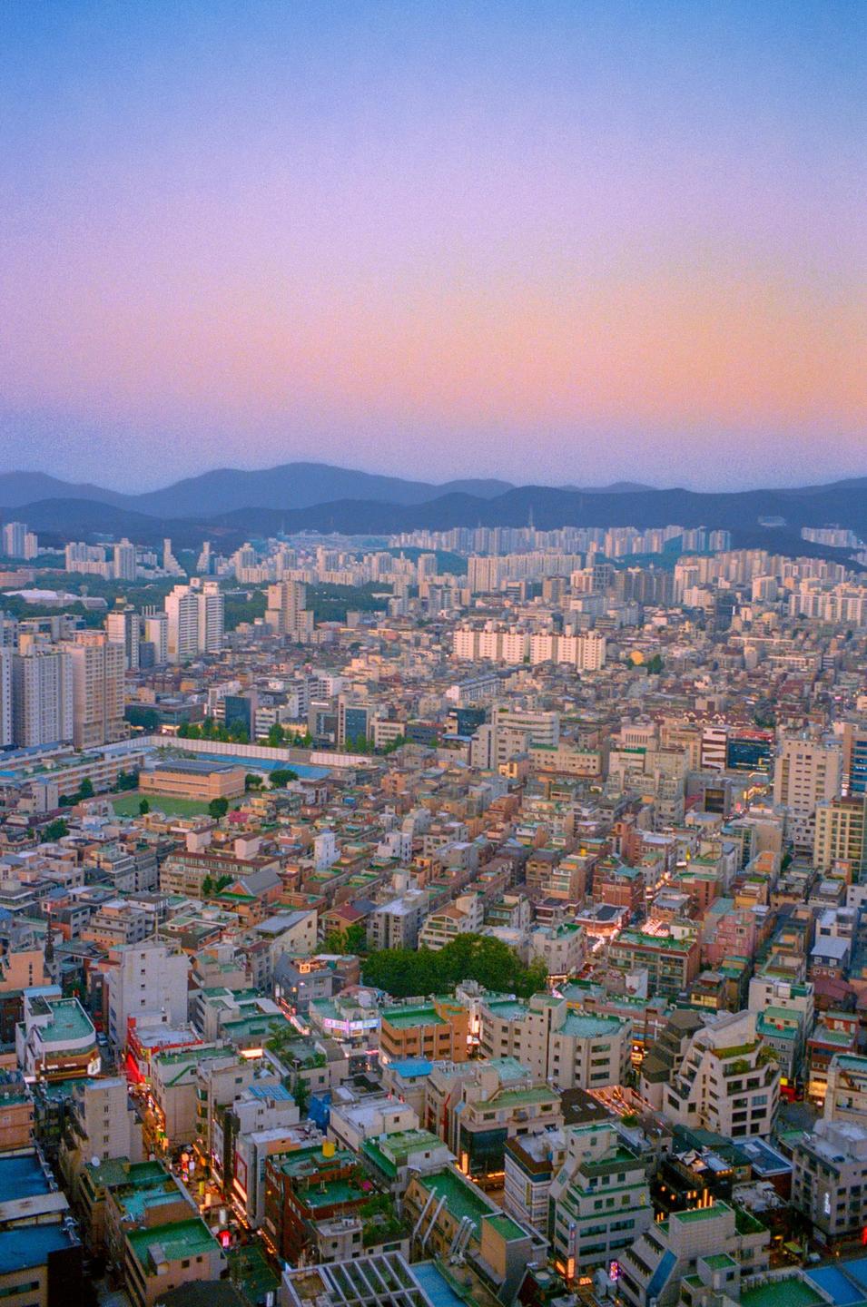 An aerial view of a city with mountains in the distance under a colorful sky at dusk.