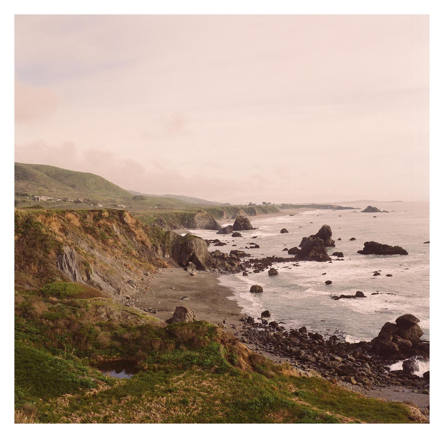 A rocky coastline with cliffs and green hills stretches along the ocean under a hazy sky.