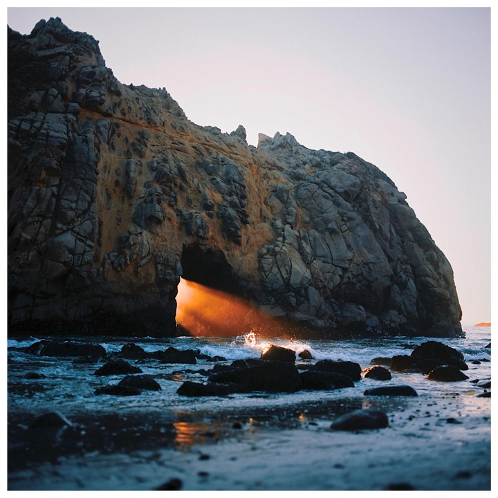 A beam of golden light shines through a natural arch in a large rock formation on a beach with waves crashing.