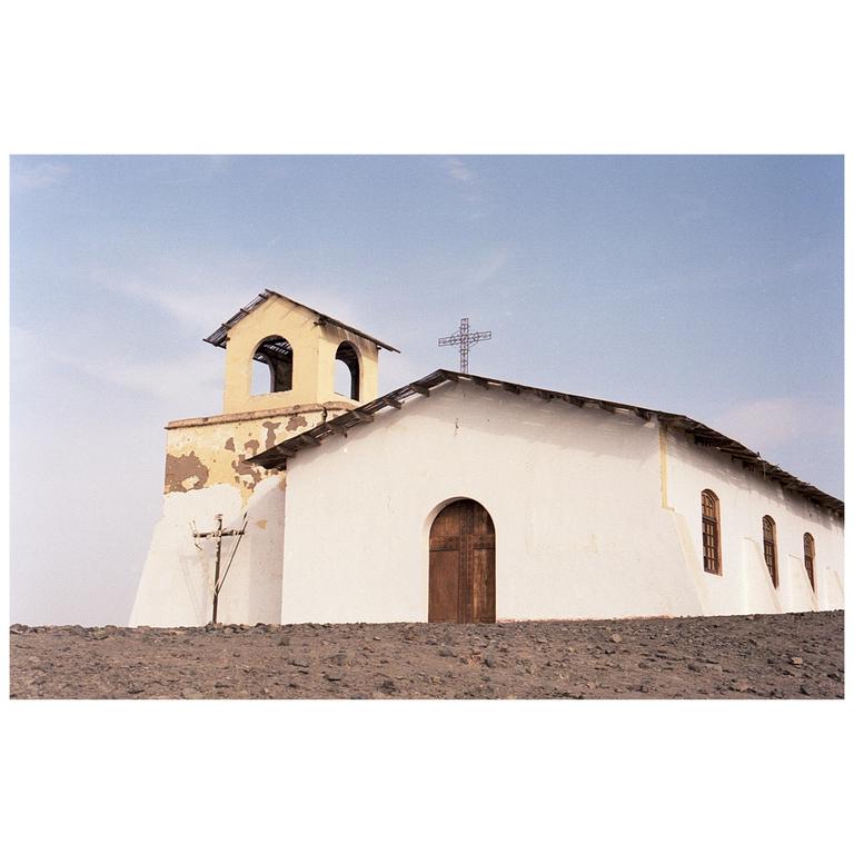 A white church with a bell tower and a cross on the roof stands on a rocky hill under a clear sky.