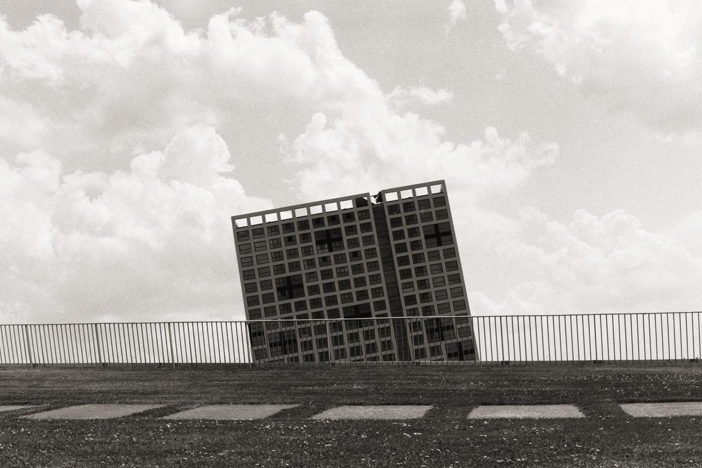 A black and white photo shows a tall building with many windows, appearing to lean to the left, behind a metal fence and a grassy area with square stepping stones.