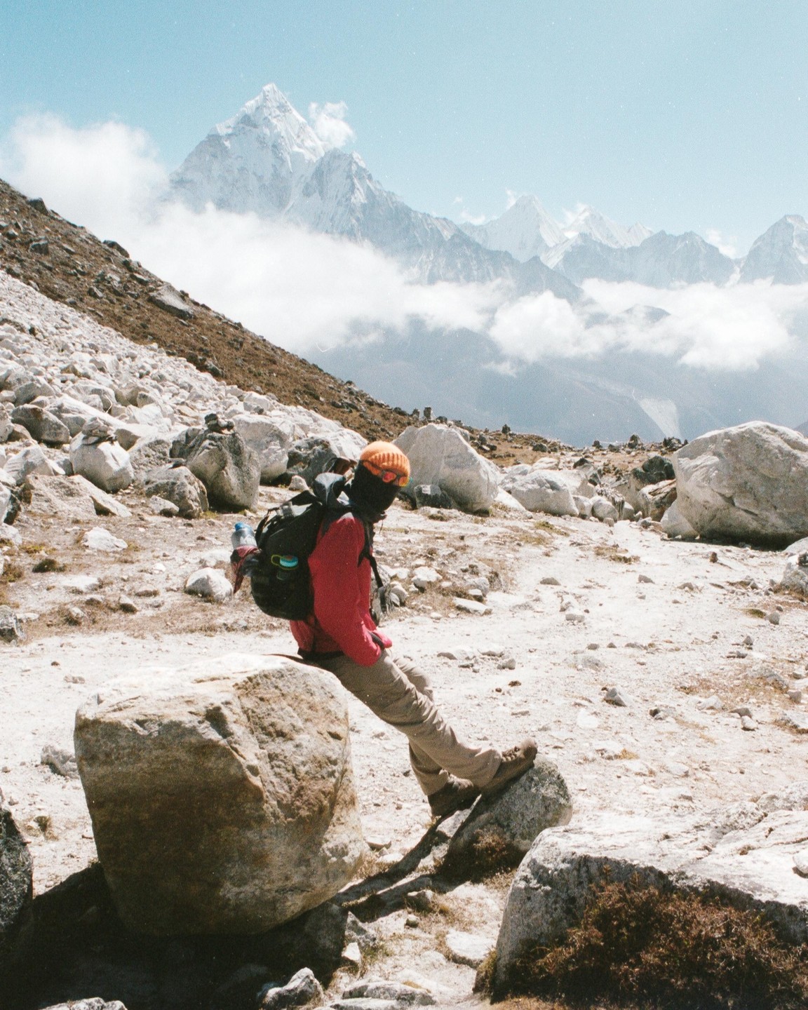 A person wearing a red jacket and an orange hat sits on a large rock in a rocky, mountainous landscape with snow-capped peaks in the background.