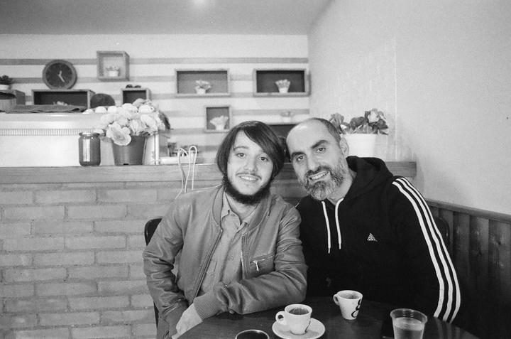 Two men with beards sit at a table with coffee cups in a cafe.