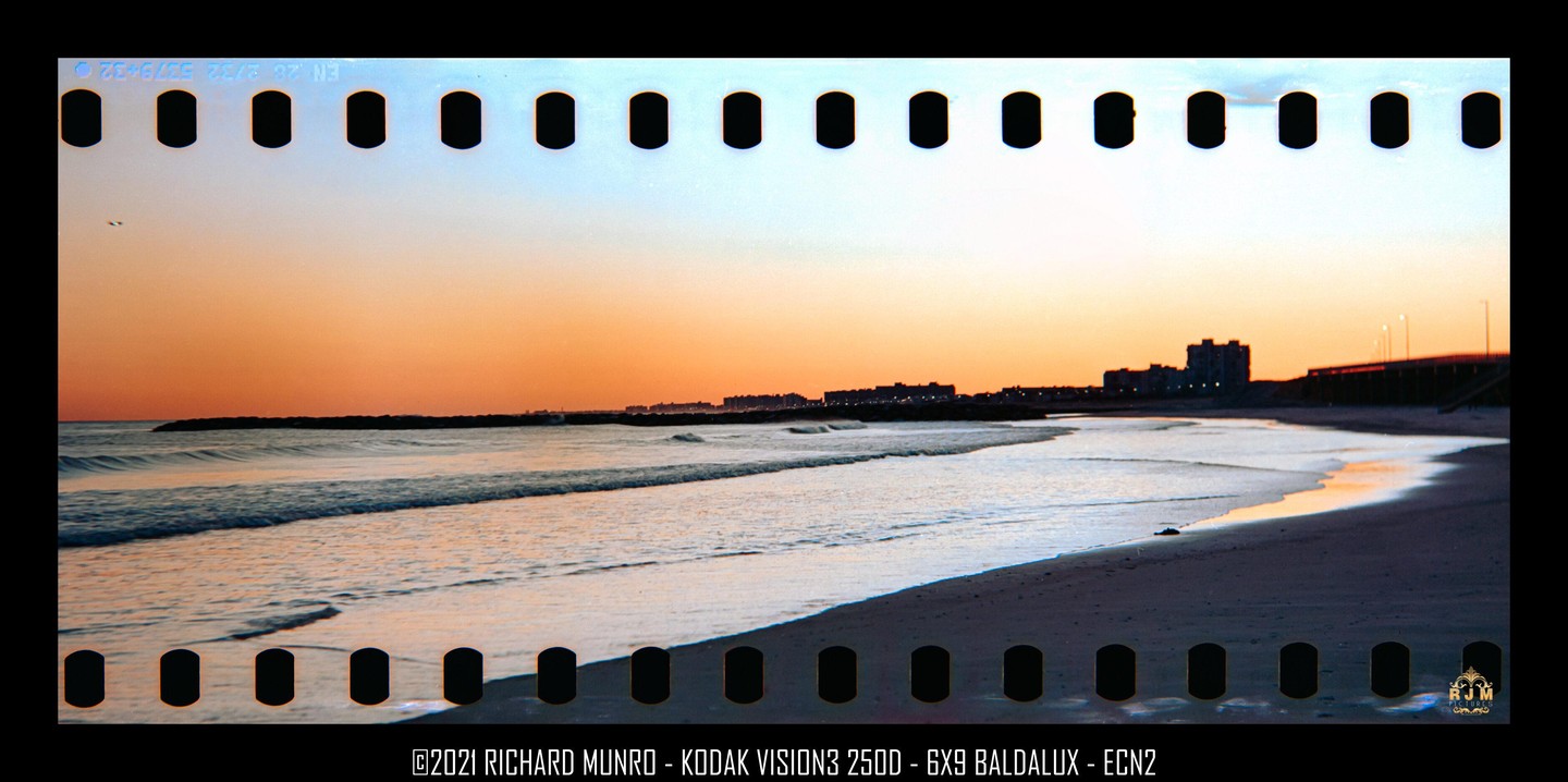 A beach with waves rolling onto the sand is visible under a colorful sky at sunset, with buildings and a bridge in the distance.