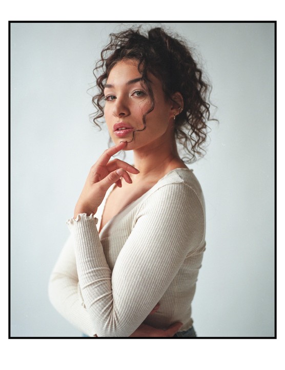 A woman with curly hair poses with her hand on her chin.