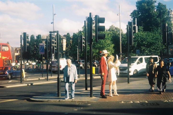 A man in a red suit and a woman in a white dress stand on a street corner with other pedestrians and vehicles.