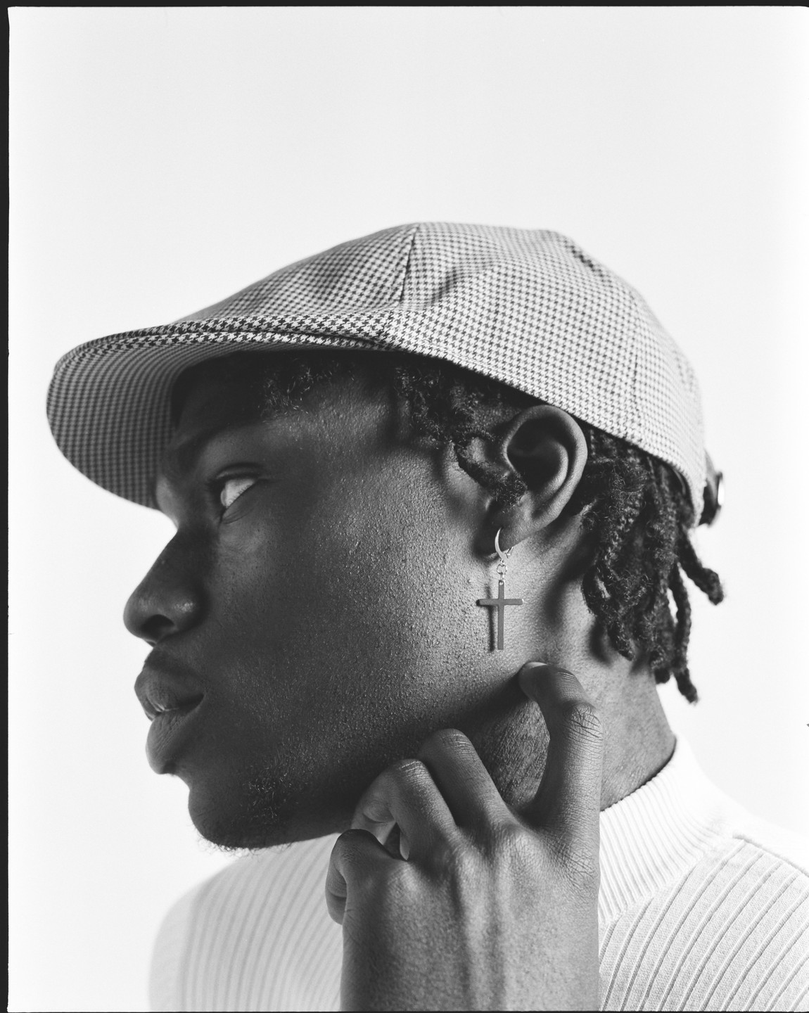 A man wearing a houndstooth flat cap and a cross earring looks up and to the right, with his hand touching his neck.