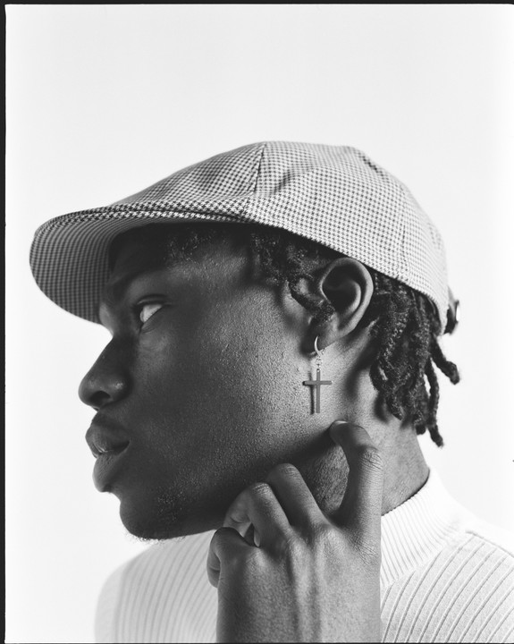 A man wearing a houndstooth flat cap and a cross earring looks up and to the right, with his hand touching his neck.
