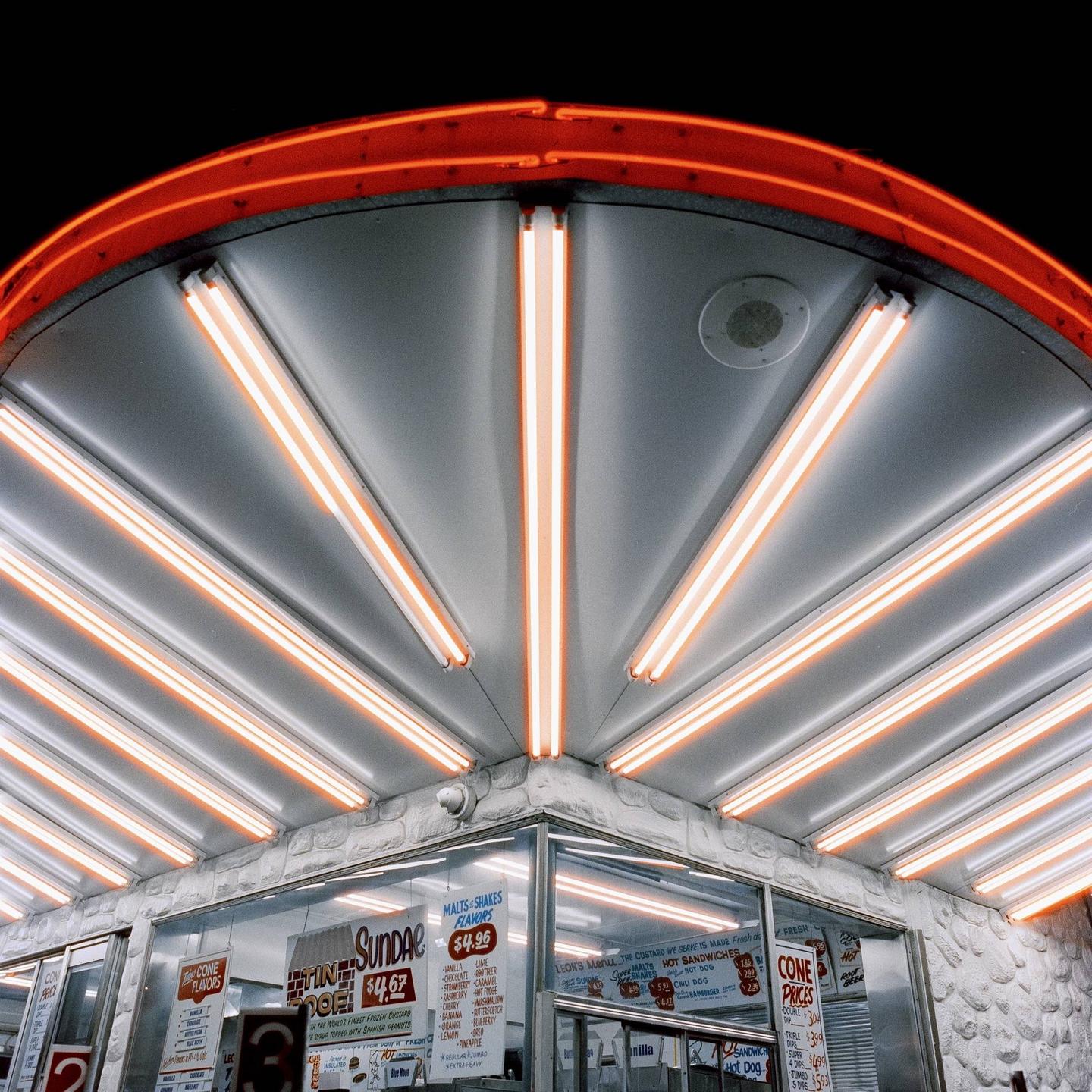 A brightly lit ice cream stand with neon lights and menu boards is shown at night.