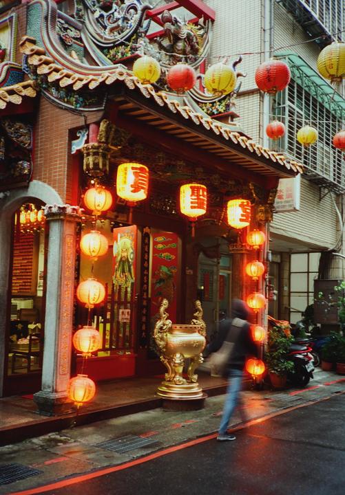 A person walks past a building adorned with many red and yellow lanterns and a large golden urn on a rainy street.
