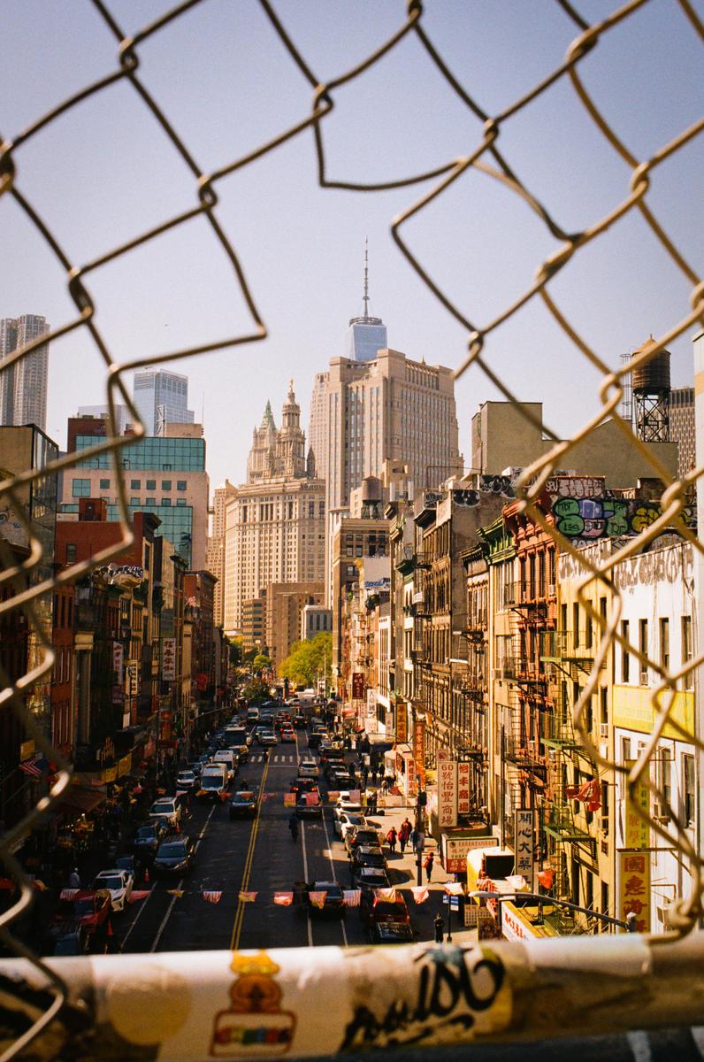 A street in a city is viewed through a chain link fence, with tall buildings and traffic visible.