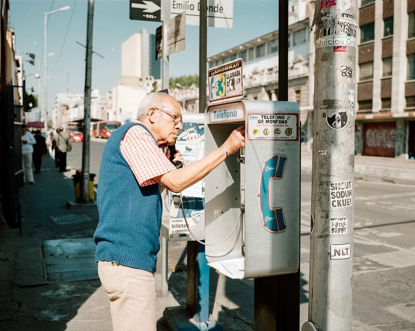 An elderly man in glasses and a blue vest is using a payphone on a city street.
