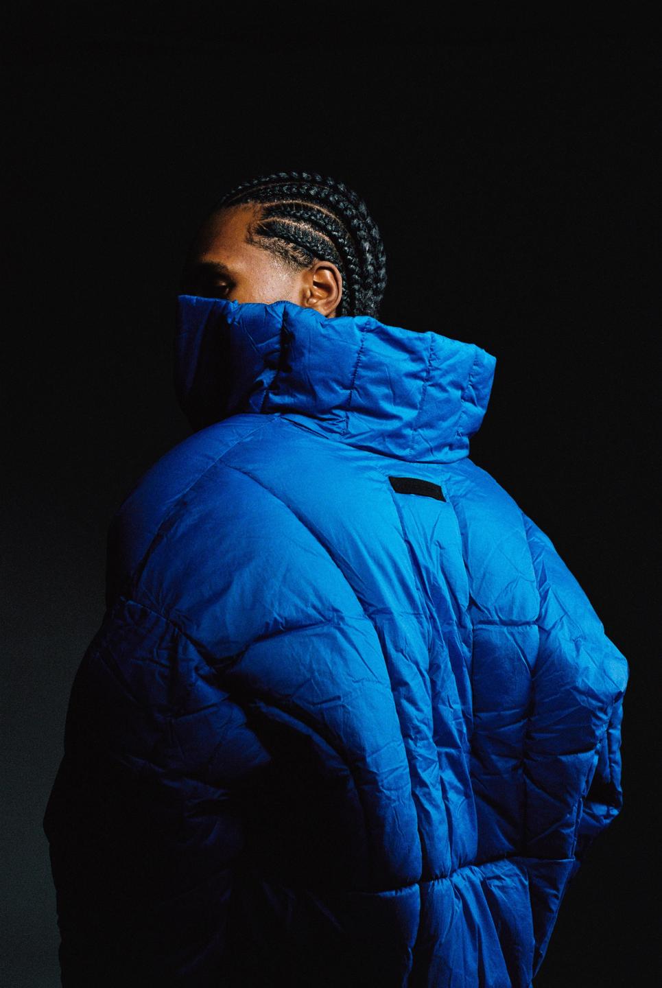 A man with braided hair wears a blue puffer jacket with a high collar that covers his face.
