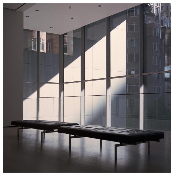 Two black benches sit on a dark wooden floor in a room with large windows looking out onto city buildings, with sunlight casting diagonal shadows across the windows.