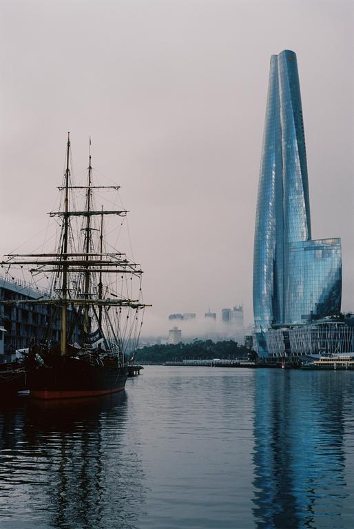 A tall ship is docked in a harbor with a modern skyscraper and a hazy city skyline in the background.
