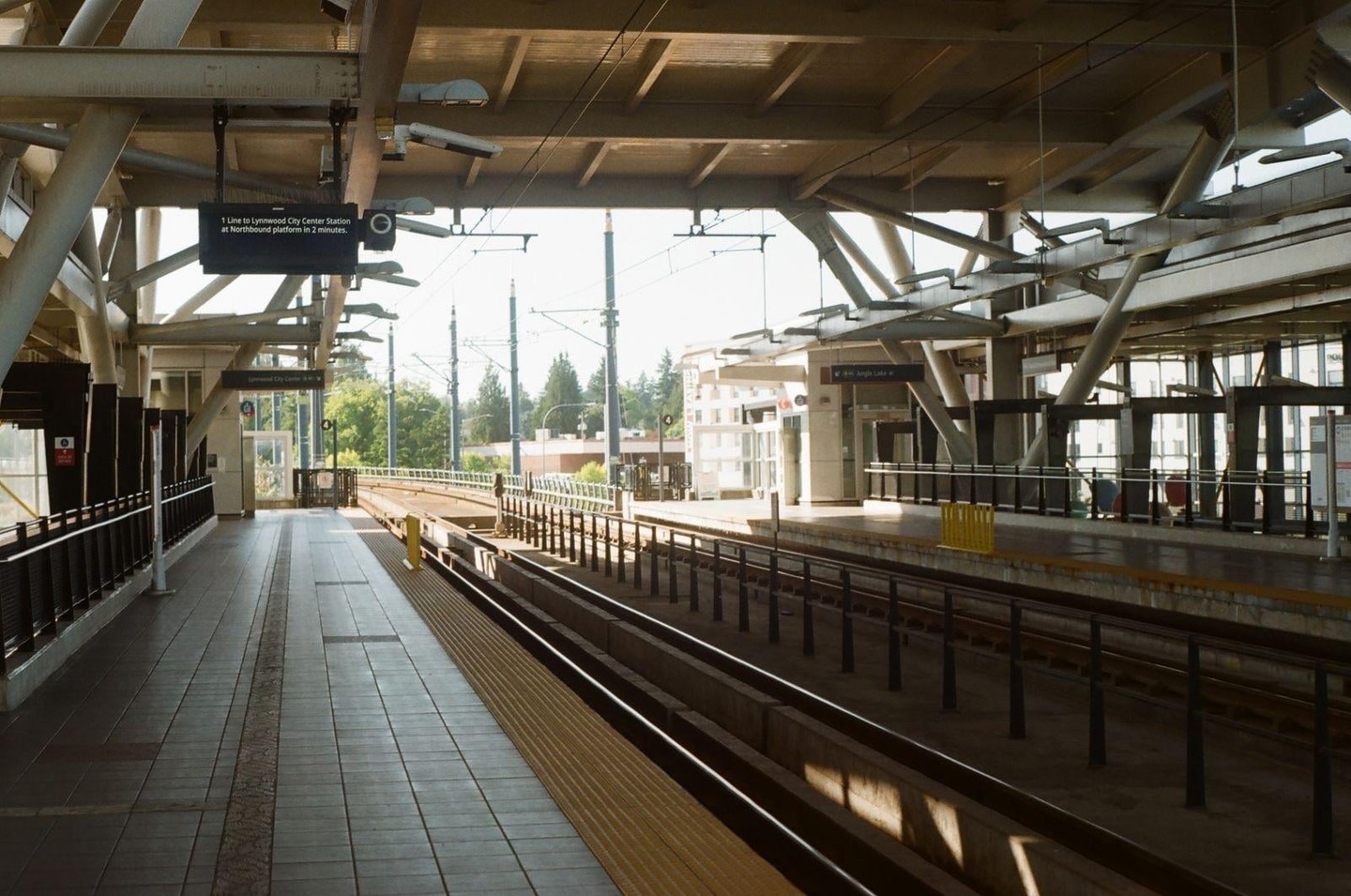 A train station platform with tracks and an overhead structure is empty under a bright sky.